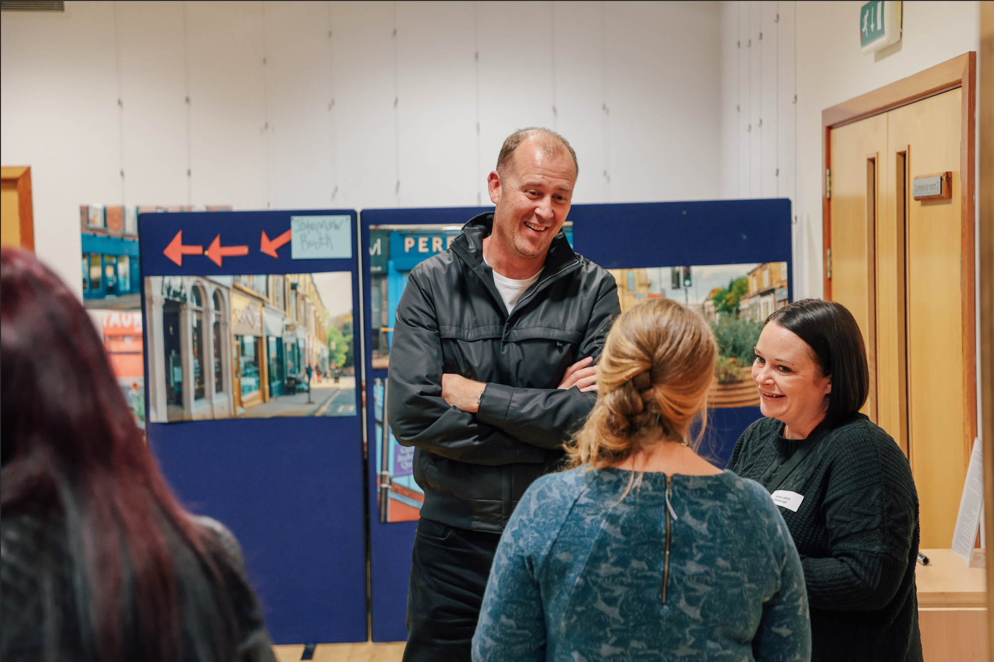 A man and a woman are engaged in conversation, surrounded by a group of people in a busy indoor setting.