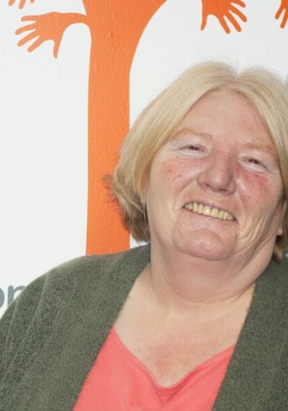 A woman with blonde hair and a green cardigan smiles brightly, showcasing a cheerful expression.