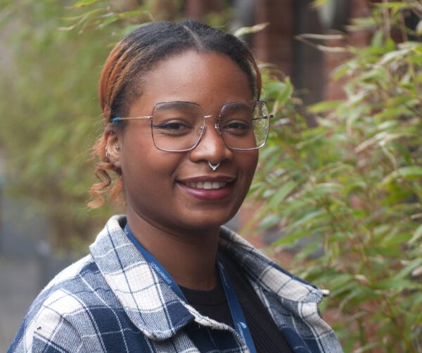 A woman wearing glasses and a plaid shirt smiles at the camera, showcasing her casual style and friendly demeanour.