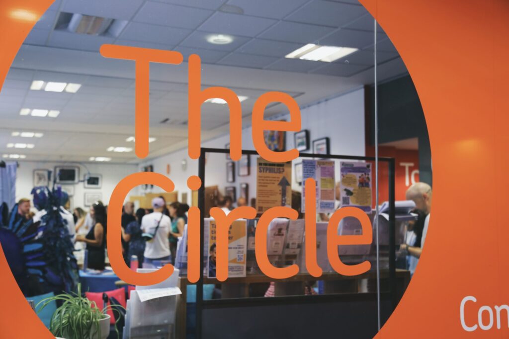 The Circle logo displayed through a indoor window through which a group of people gathering in the Gallery can be seen