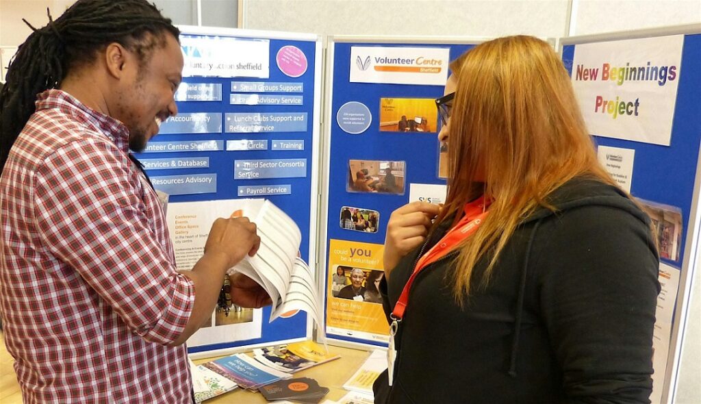 Two people are standing in front of a blue board with information about volunteering.